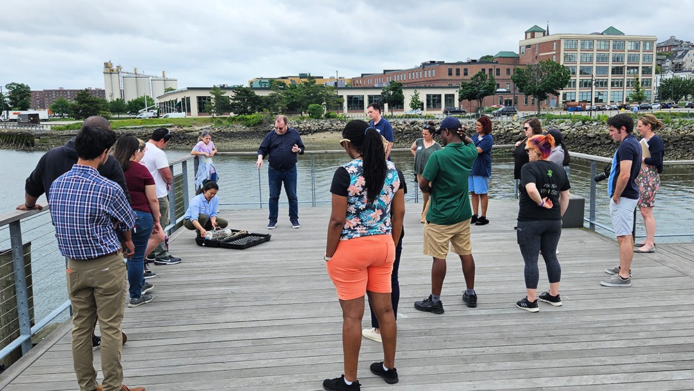 Haley & Aldrich STEM Learning Day for Boston Public School District. Group of people standing on a deck near a body of water watching Haley & Aldrich representative.
