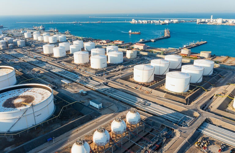 An aerial shot of an industrial port shows a large number of white storage tanks, likely for oil or chemicals, clustered along the coastline. Some tanks are significantly larger than others, and one in the foreground has a large, rusty stain on its top. Numerous pipes and access roads connect the tanks, leading toward a calm blue sea, where several ships are visible in the distance. The shoreline stretches out to the horizon, dotted with more industrial structures and what appears to be part of a city or port infrastructure. 