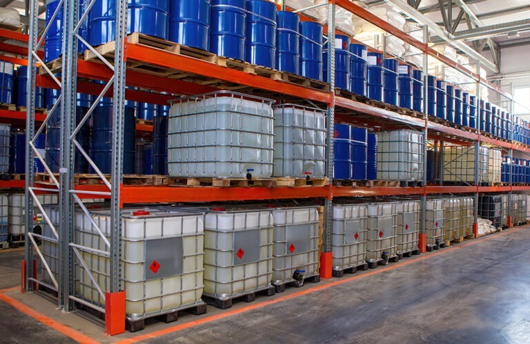A wide shot shows a large warehouse with several rows of tall metal shelving units filled with various containers. The top shelves hold numerous blue barrels, while the lower shelves are stacked with large, square, clear, or translucent plastic totes, many of which have red diamond-shaped hazard symbols. 