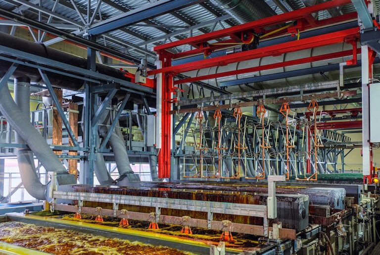 A large industrial interior shows a metal plating operation with several tanks containing an orange, frothy liquid in the foreground. Overhead, a red gantry crane system holds numerous metal racks with hooks ready to immerse items in the tanks for plating. The background features complex industrial machinery, including large pipes and a metal ceiling structure, all under bright lighting. 