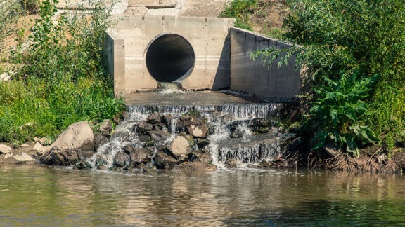 A concrete drainage pipe empties into a rocky stream, creating a small waterfall. Green foliage grows on the banks of the stream, and the water in the foreground reflects the sky.