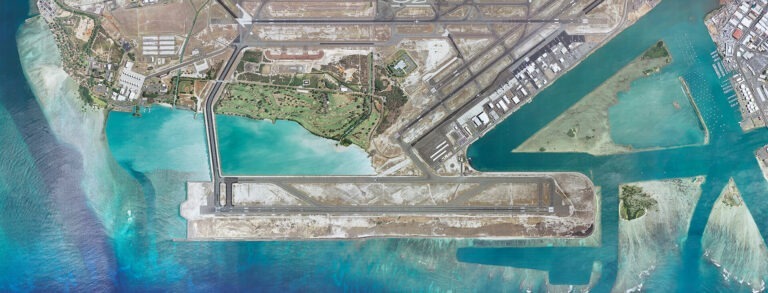 Aerial view of Daniel K. Inouye International Airport with multiple runways and taxiways extending onto artificial land surrounded by clear blue ocean water.