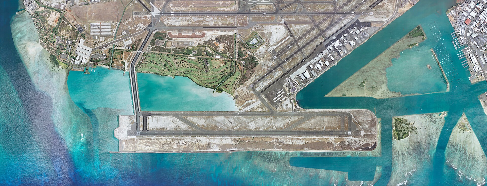 Aerial view of Daniel K. Inouye International Airport with multiple runways and taxiways extending onto artificial land surrounded by clear blue ocean water.