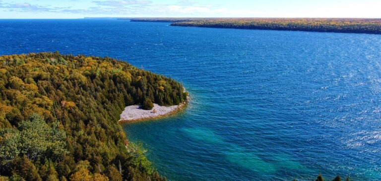 Aerial view of a forested shoreline and rocky beach along a Great Lake with blue water and a distant tree-lined coast.