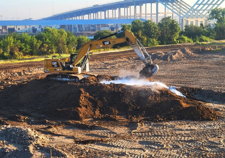 Yellow Caterpillar excavator digging into a mound of soil with visible steam or vapor rising, at a construction site with cleared land and tire tracks.