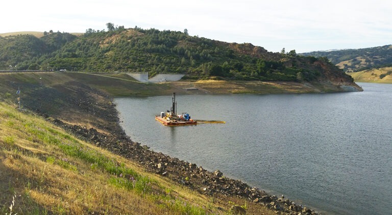 A barge-mounted drill rig is positioned on a reservoir near a rocky embankment with hills and trees in the background.