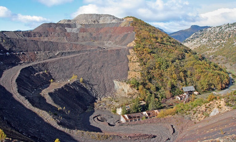 A wide view of a terraced open-pit mine beside a forested hillside, with winding dirt roads and a few abandoned buildings at the bottom.