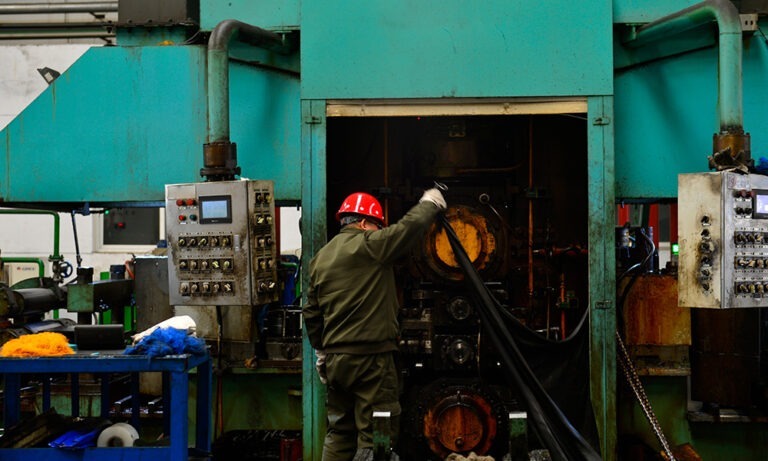 A worker wearing a red hard hat operates a large industrial machine, pulling material from its opening inside a factory setting.