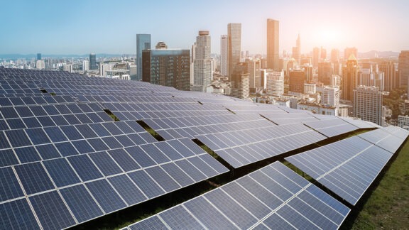 A large field of solar panels stretches across the foreground with a modern city skyline and a bright sunrise in the background.