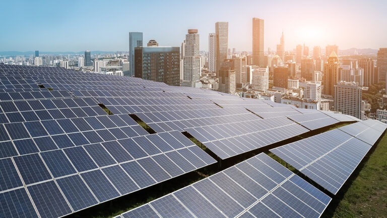 A large field of solar panels stretches across the foreground with a modern city skyline and a bright sunrise in the background.