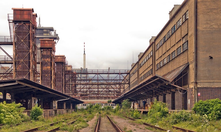 Abandoned industrial rail yard between old factory buildings with overgrown train tracks and rusted metal structures.