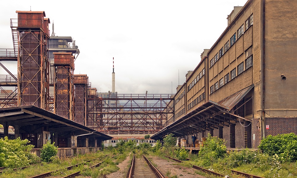 Abandoned industrial rail yard between old factory buildings with overgrown train tracks and rusted metal structures.