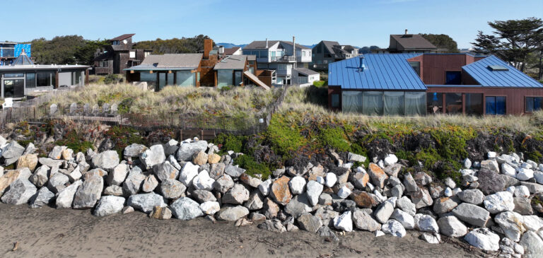 Homes behind dunes and a rock seawall on the coast.