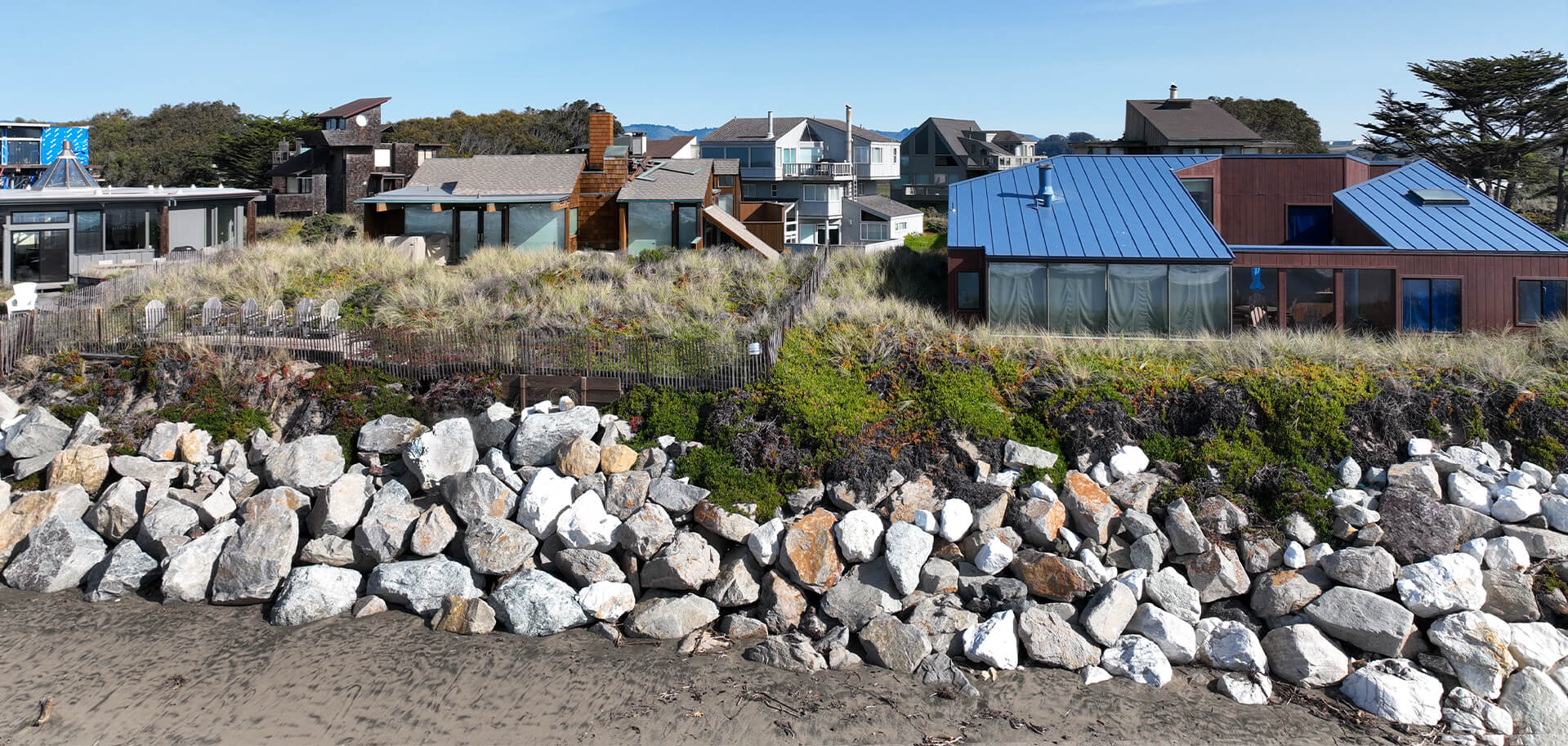 Homes behind dunes and a rock seawall on the coast.