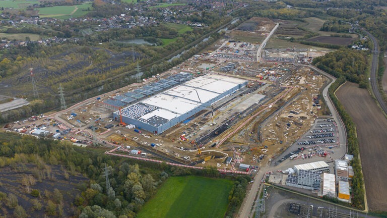An aerial view of a large industrial construction site with multiple buildings, cranes, and surrounding roads in a semi-rural area.