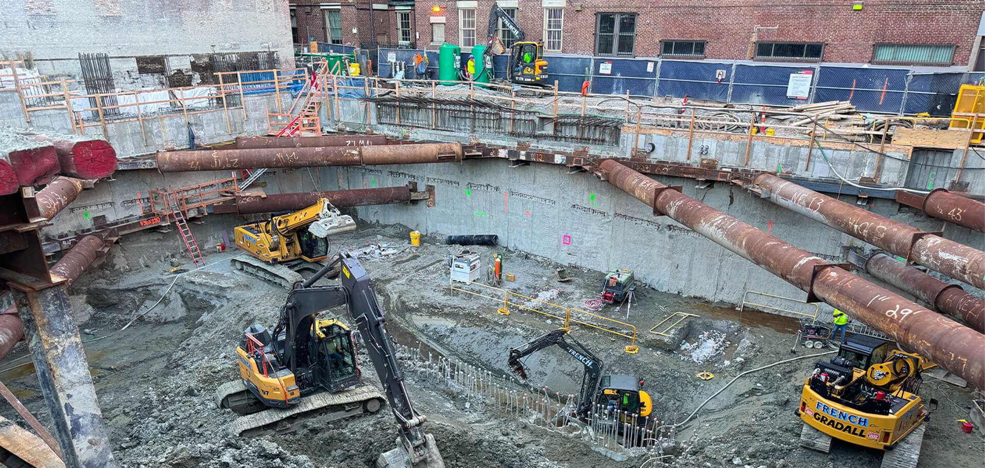 Construction workers and heavy equipment operate in a deep excavation supported by large steel cross braces and concrete retaining walls.