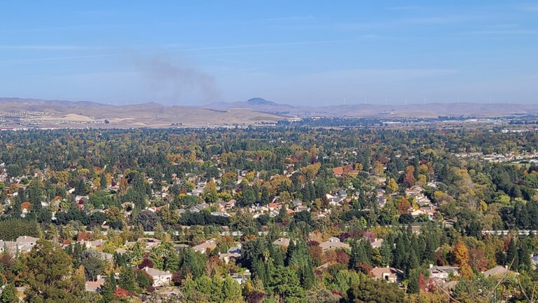 Smoke rises beyond a suburban neighborhood near dry hills and wind turbines.